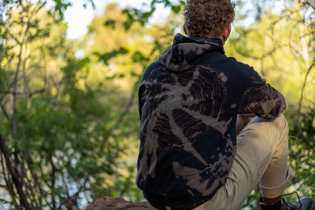 Person sitting on a log in a forest, wearing a tie-dye hoodie and beige pants.